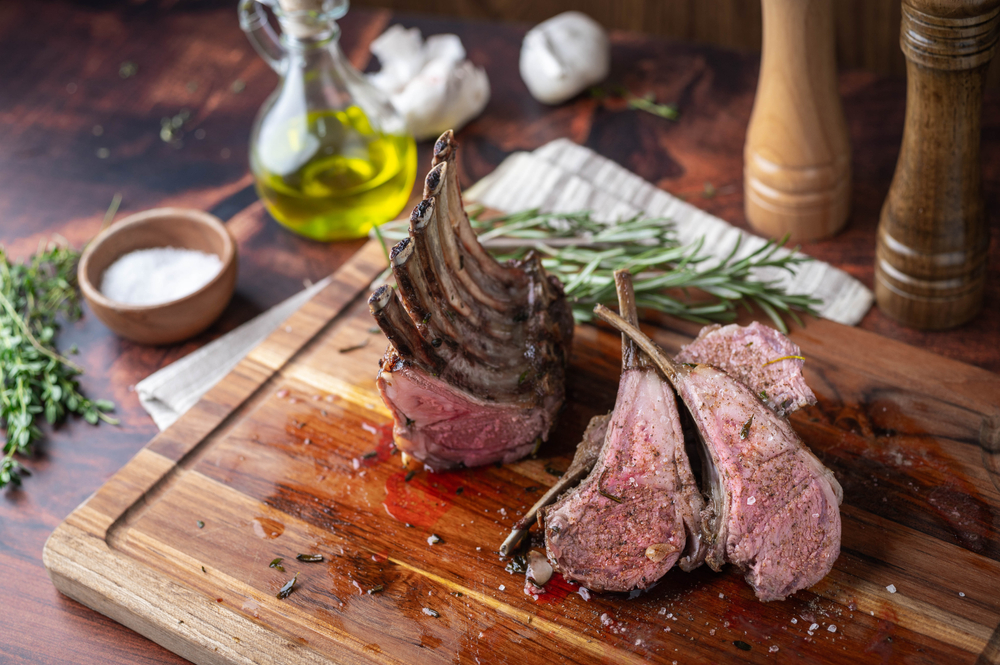 lamb rack on wood cutting board surrounded by herbs and spices