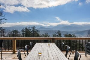 Cozy wooden deck with a large pine table surrounded by chairs of varying designs. One chair near the edge overlooks Great Smoky mountain in the distance. On the table, two glasses of beer invite relaxation and enjoyment of the view. The scene exudes a peaceful atmosphere, ideal for a mountain retreat or cabin getaway.