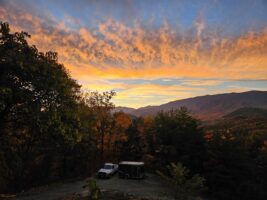 A black truck with a trailer parked on a dirt road in the mountains during the evening. Nearby, a van and an SUV are also parked. The sun sets behind the mountain range, painting the sky in shades of orange and yellow, creating a beautiful sunset. Tall pine trees and a forest surround the vehicles, adding to the picturesque mountain landscape and enhancing the tranquility of the setting.