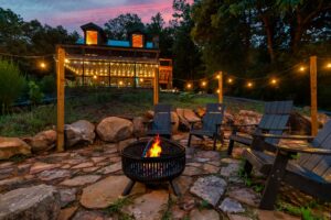 Cozy outdoor setting with a fire pit surrounded by black plastic chairs, illuminated by lights. Candles in the fire pit add to the warm and inviting ambiance. The scene includes a stone patio and a view of pine trees and mountains in the distance, creating a serene atmosphere perfect for a relaxing evening outdoors.